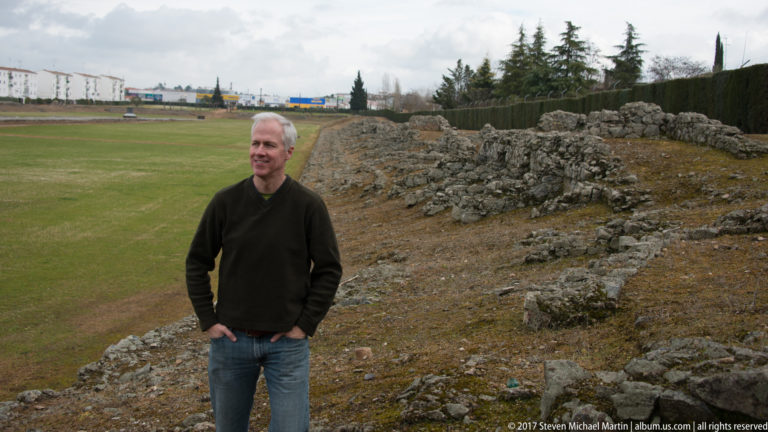 Man standing beside rough stone archaeological remains with the grassy interior of an ancient amphitheatre behind him.