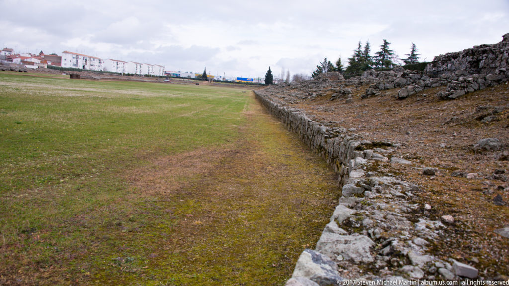Long low stone wall bordering the grassy interior of Mérida's Roman amphitheatre, with modern buildings in the distance.