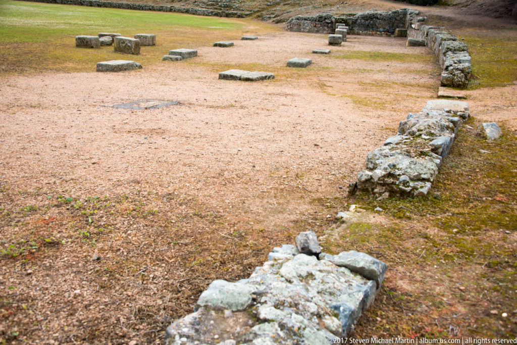 Low stone foundations and scattered blocks outlining a rectangular area within the archaeological amphitheatre complex.