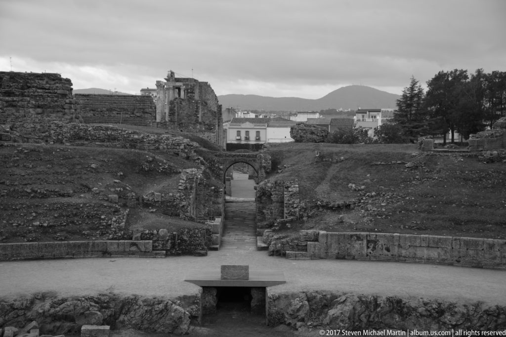 Black-and-white view down a straight passage between archaeological walls toward an arched opening and distant ruins.