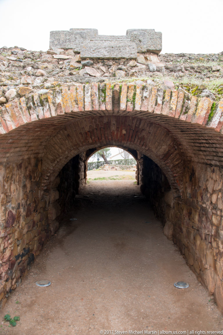Low brick barrel vault over a stone-walled passage, with a second arch and daylight visible at the far end.