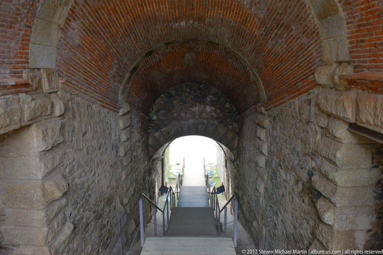 Stairway descending through a tall brick-and-stone vaulted passage with modern handrails beneath the amphitheatre seating.