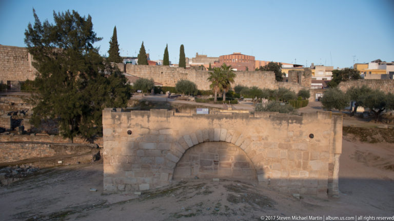 Low rectangular stone structure with a broad round arch, surrounded by archaeological ground and olive trees.