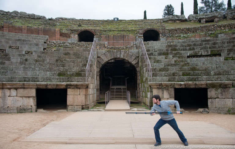 Man holding a long monopod across the amphitheatre arena in front of a central arched entrance and stone seating.