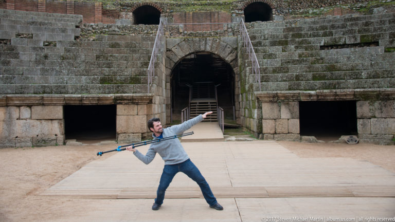 Man striking a playful pose with a long monopod before the central entrance and stone seating of the Roman amphitheatre.