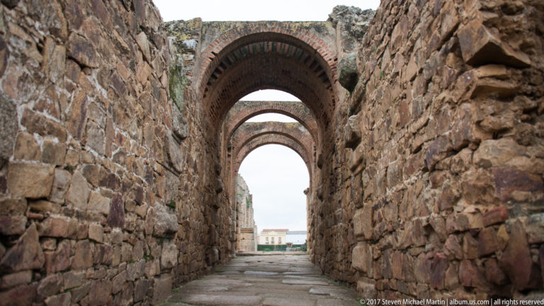 Narrow stone corridor crossed by several receding brick arches and opening to daylight at the far end.