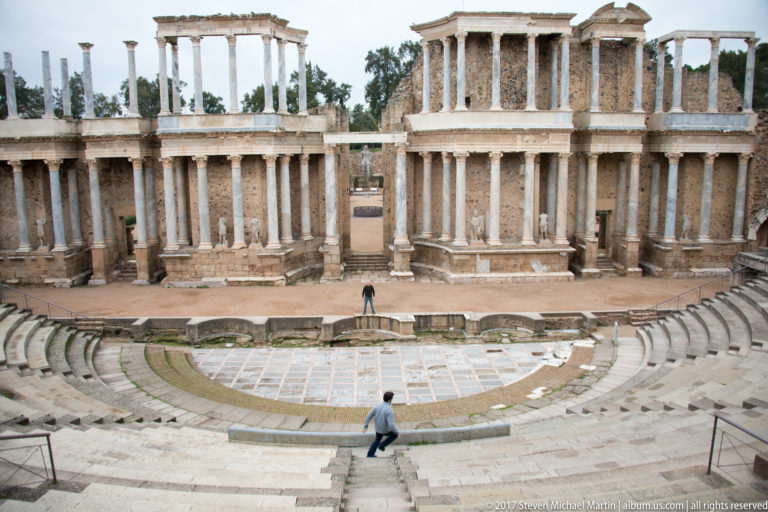 Roman Theatre stage façade beyond the orchestra, with visitors positioned on the stage area and descending the stone seating.