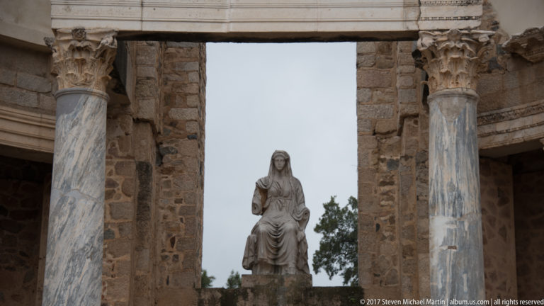 Seated draped female statue positioned between tall marble columns on the stage façade of Mérida's Roman Theatre.