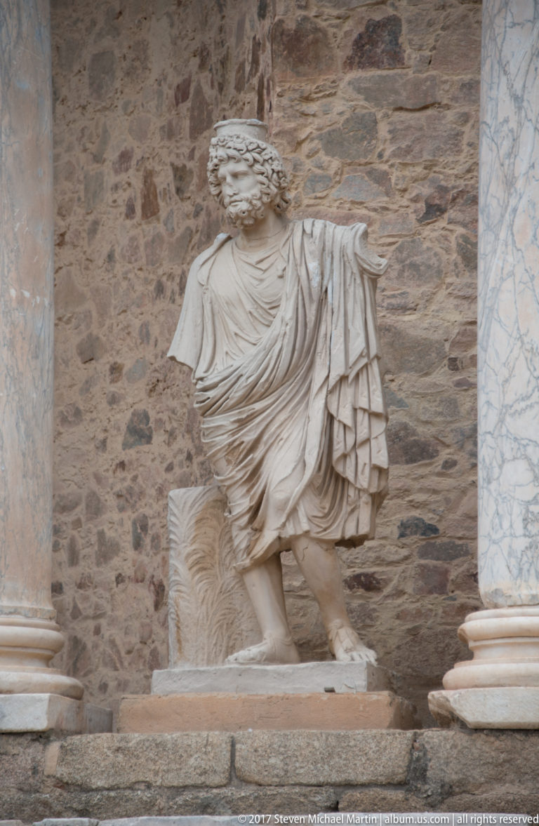 Headless draped male marble statue standing in a niche against rough stone masonry on the Roman Theatre stage façade.