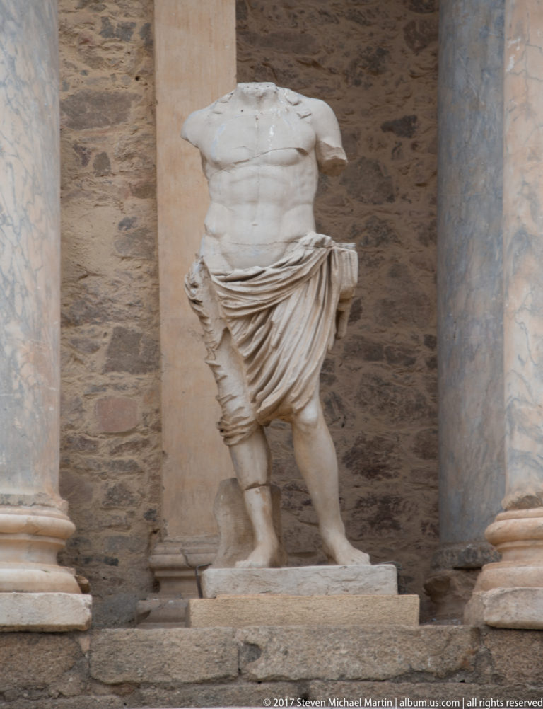 Headless marble male statue with muscular torso and deeply carved drapery, positioned between columns on the theatre stage façade.