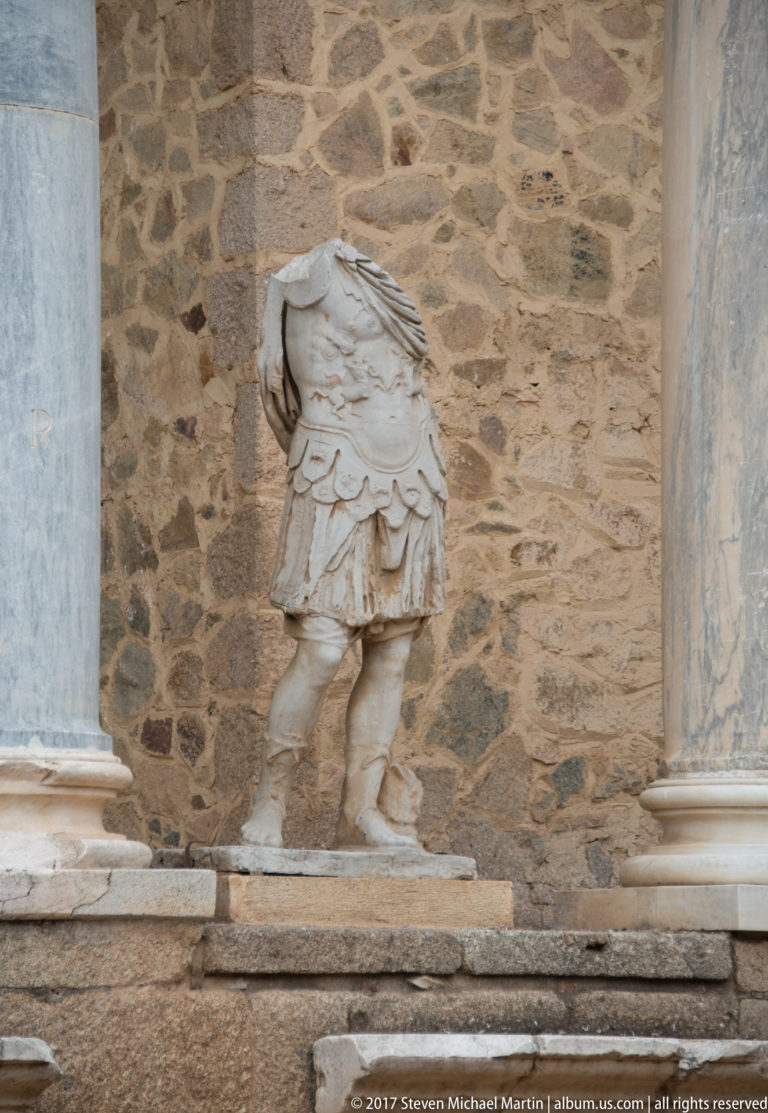 Headless marble male statue wearing a short armored garment, standing between columns against the theatre's stone stage wall.