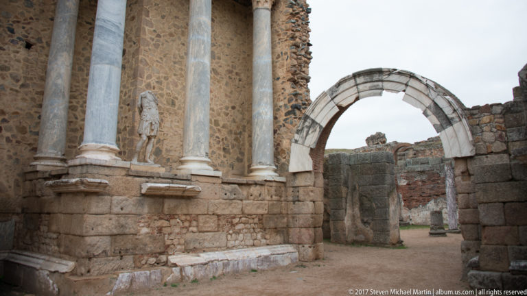 Marble columns and statue beside a large stone arch in a side section of Mérida's Roman Theatre.