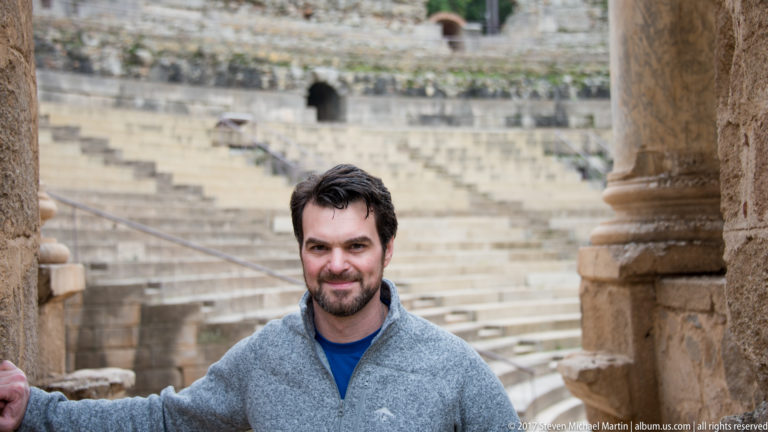 Bearded man in gray fleece posing beside a stone column, with the Roman Theatre seating blurred behind him.