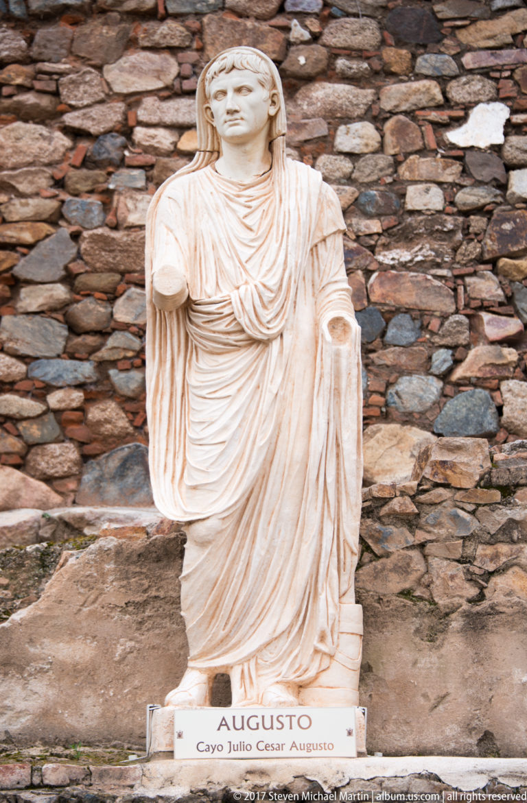 Standing pale statue of Augustus in Roman drapery before a rough stone wall, with a label naming Cayo Julio César Augusto.
