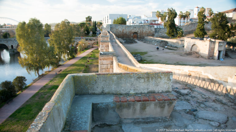 Curving plastered parapet on an elevated fortification, with long wall, trees, path, and river beyond.