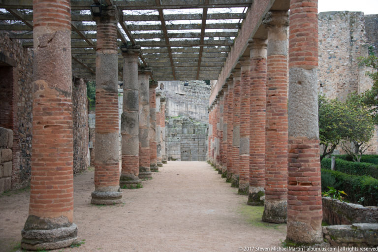 Long columned portico with brick piers, stone columns, overhead framework, and garden areas behind Mérida's Roman Theatre.