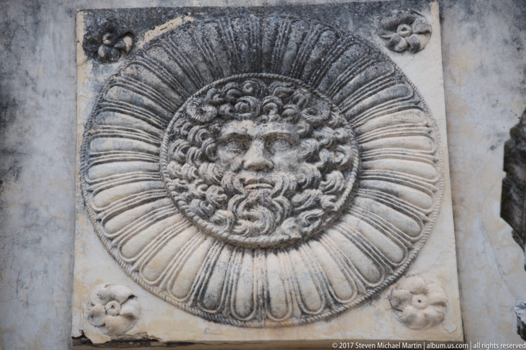 Close view of a bearded face carved in a circular radiating relief with small rosettes at the corners.