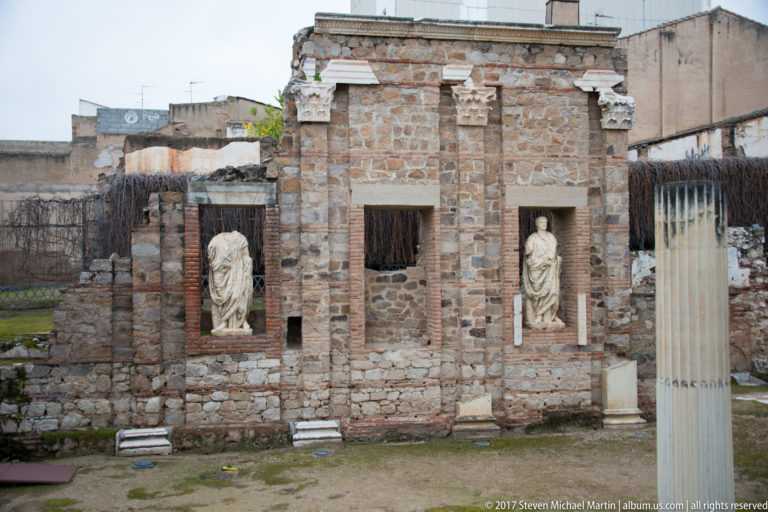Stone-and-brick forum wall with two draped statues in tall niches and a central rectangular opening.