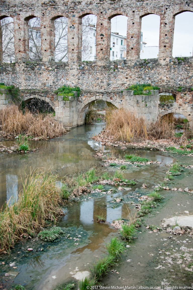 Lower aqueduct arch crossing a shallow stream amid reeds, with the taller Roman arcade rising above.