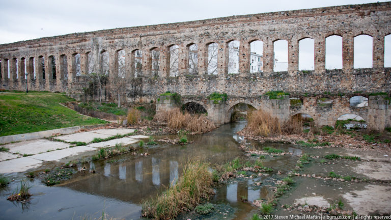 Roman aqueduct arches above shallow water, reeds, and grassy banks in Mérida's Albarregas valley.