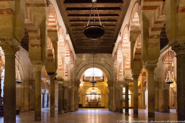 Striped arches and stone columns form an aisle ending at a brightly lit Christian devotional area inside the Mezquita.