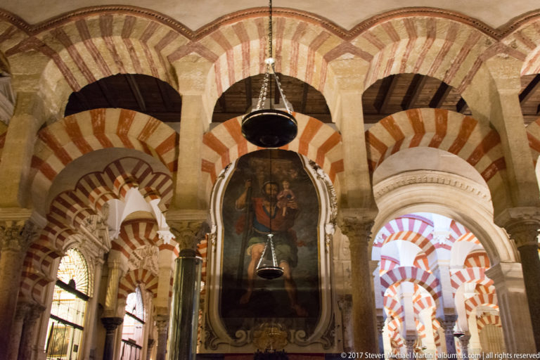 Large framed Christian religious painting hanging among striped arches, stone columns, and suspended lamps inside the Mezquita.