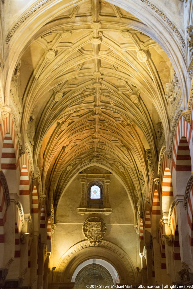 Ornate elongated stone vault with a hanging chandelier, bordered by striped mosque arches inside the Mezquita.