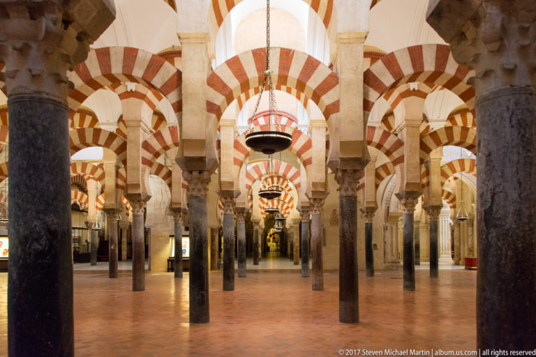Wide interior view across dark columns, suspended lamps, and repeating red-and-light striped arches in the Mezquita.