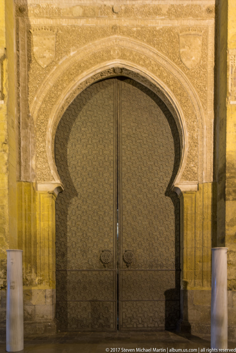 Decorated horseshoe-arched doorway with patterned metal double doors set into the stone exterior wall of the Mezquita.