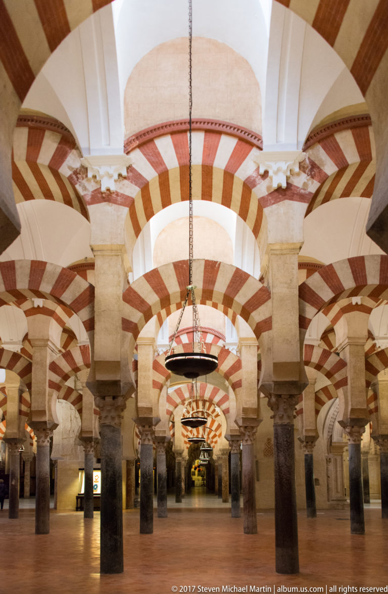 Low symmetrical view down repeated striped arches and varied stone columns, with hanging lamps aligned along the center.