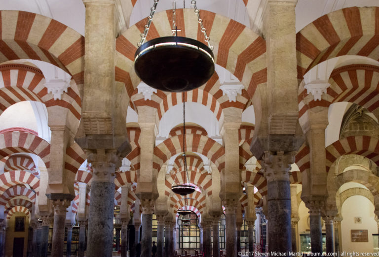 Close interior view through striped double arches and stone columns with large suspended lamps in the Mezquita.