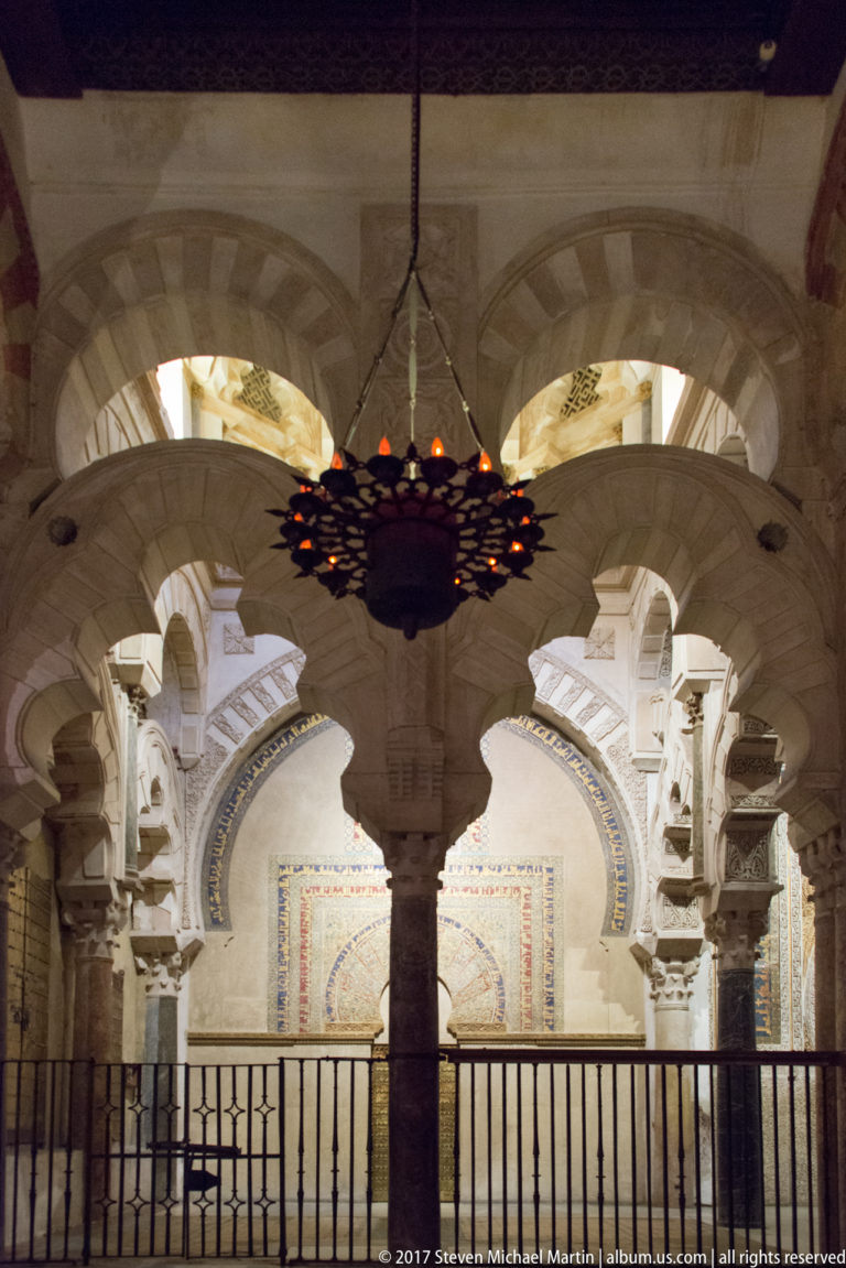 Ornate pale lobed arches and carved details seen behind a metal screen and hanging lamp inside the Mezquita.