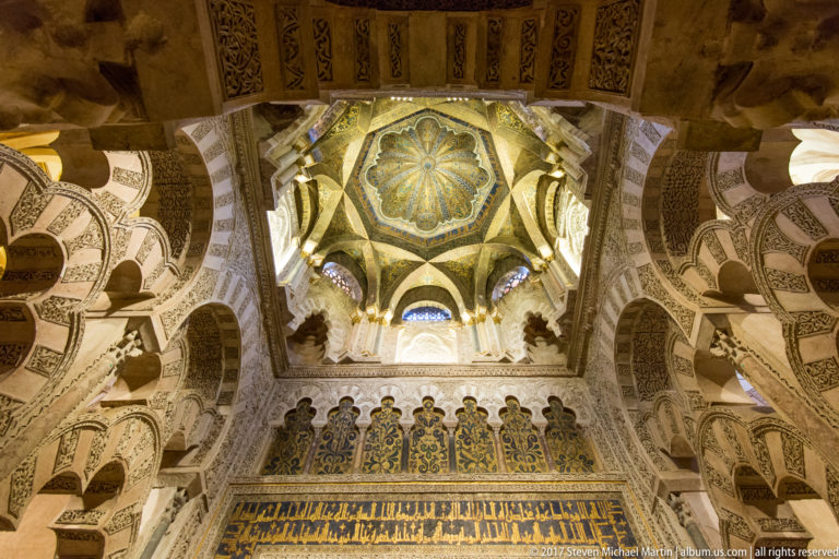 Upward view of the ornate maqsura dome, intersecting lobed arches, and patterned surfaces above the Mezquita mihrab.