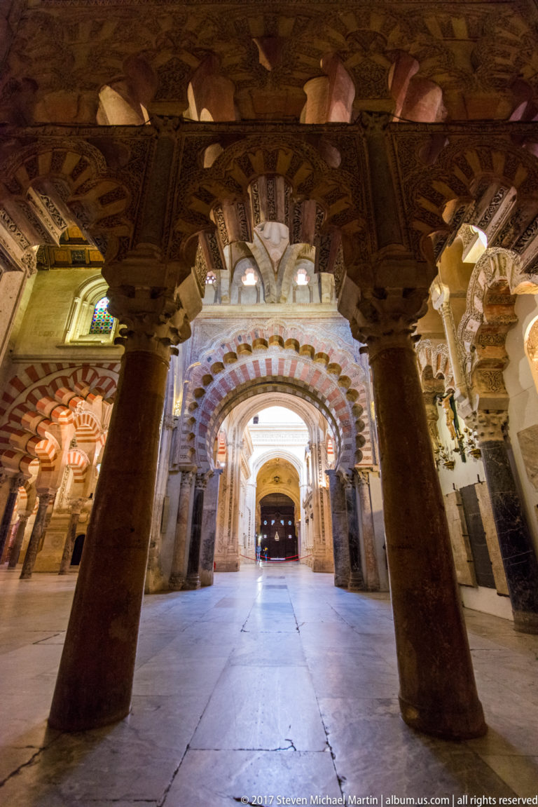 Low-angle interior view between dark columns toward a bright red-and-light striped arch in the Mezquita.