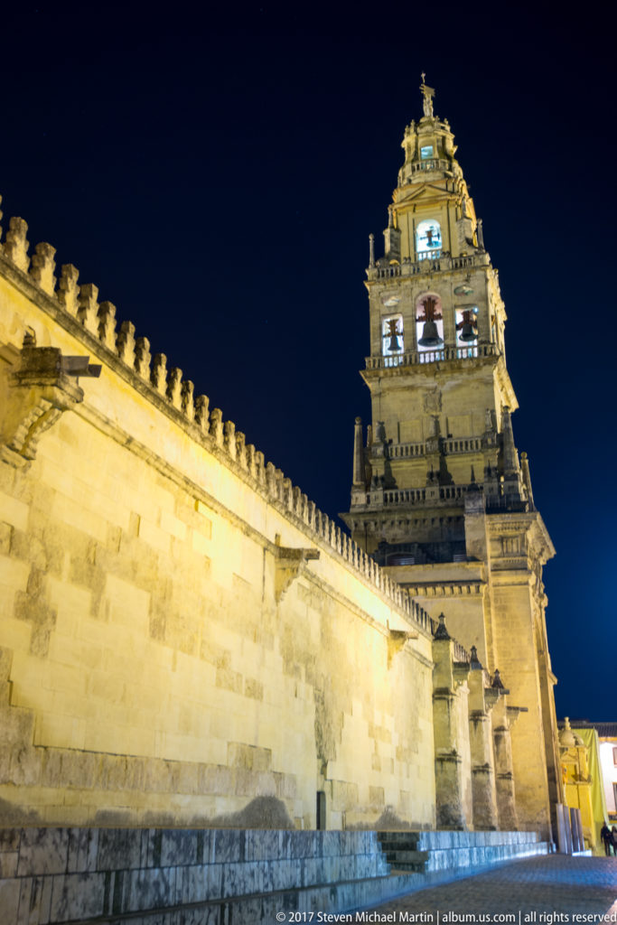 Crenellated stone wall of the Mezquita leading toward its illuminated bell tower under a deep blue night sky.