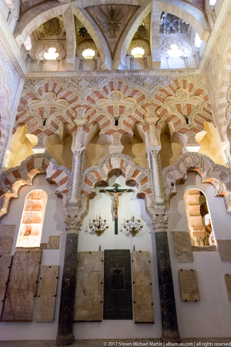 Intersecting multilobed striped arches above slender columns, with a crucifix and wall plaques below inside the Mezquita.