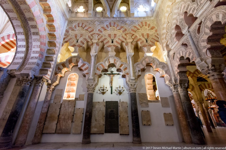 Multilobed red-and-light arches rise above a crucifix and rectangular wall plaques inside the Mezquita.