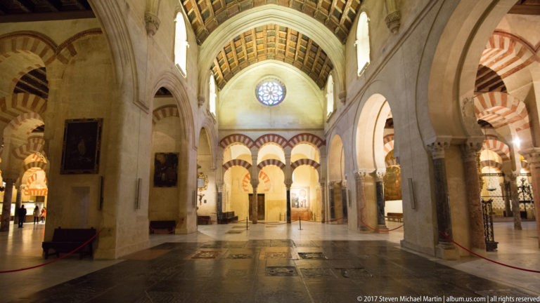 Tall cathedral nave with pale piers, paneled ceiling, rose window, and striped arches visible at the far end.