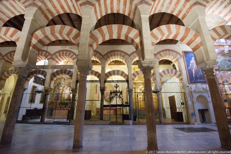 Three striped arches and columns frame a metal-screened chapel with a crucifix inside the Mezquita.
