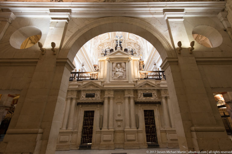 Pale Renaissance-style cathedral enclosure with pilasters, barred openings, relief sculpture, and balustrade, framed by a large arch.