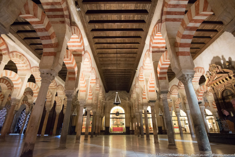 Long aisle beneath a dark timber ceiling, lined by striped arches, stone columns, and hanging lamps in the Mezquita.