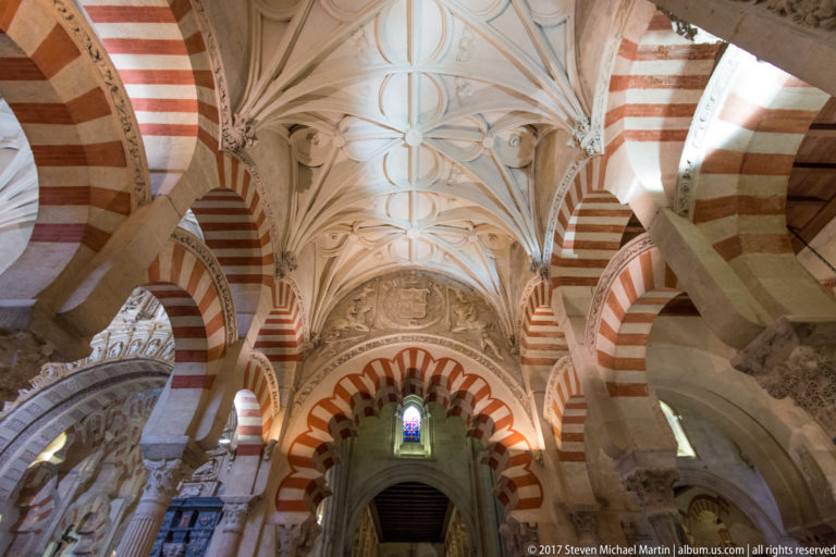 White ribbed vault above striped arches and carved stone supports, with a small stained-glass window at center.