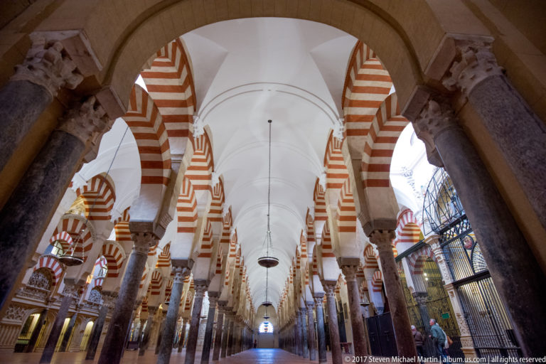 Symmetrical Mezquita aisle lined with dark columns, striped arches, pale vaulting, and suspended lamps.