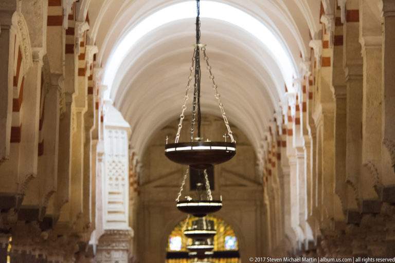 Large circular metal lamps hang in a receding line down a softly focused Mezquita aisle.
