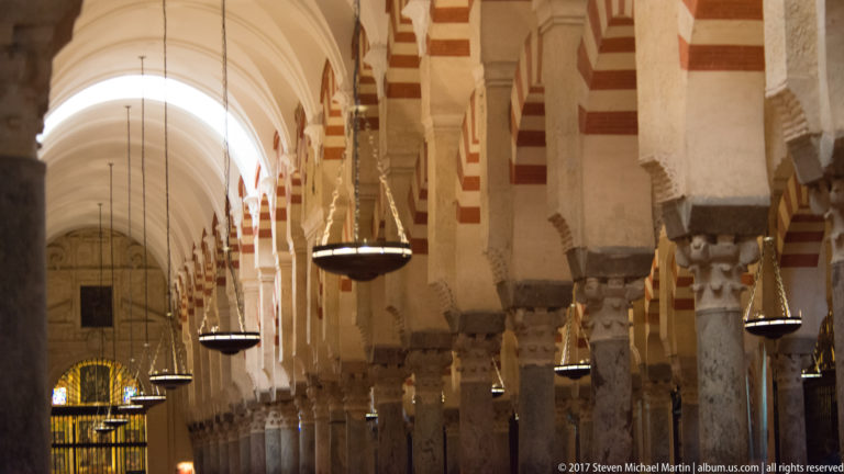 Oblique interior aisle with hanging lamps, massive piers, striped arches, and a bright stained-glass opening in the distance.