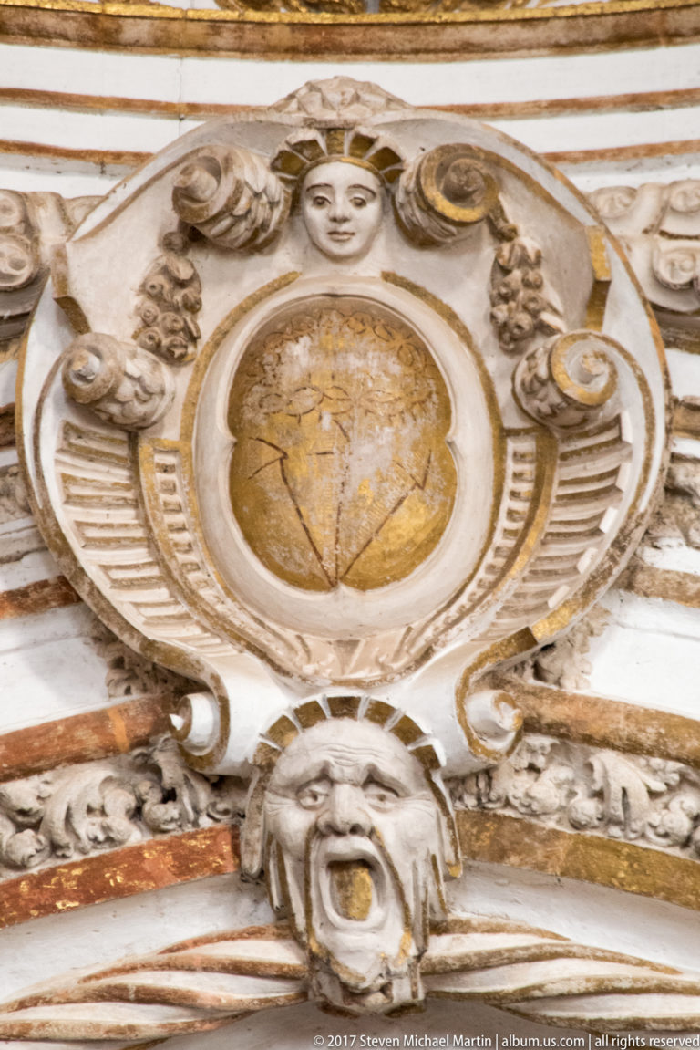 Baroque decorative medallion with central roundel, scrollwork, small faces, and a mask-like head below.