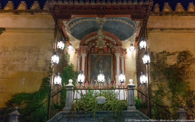 Exterior devotional shrine with a sacred image, hanging lamps, iron railings, and greenery against the Mezquita wall.