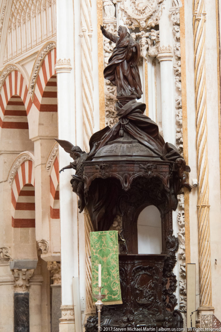 Ornately carved dark wooden pulpit with sculptural canopy, green textile, and striped Mezquita arches behind.