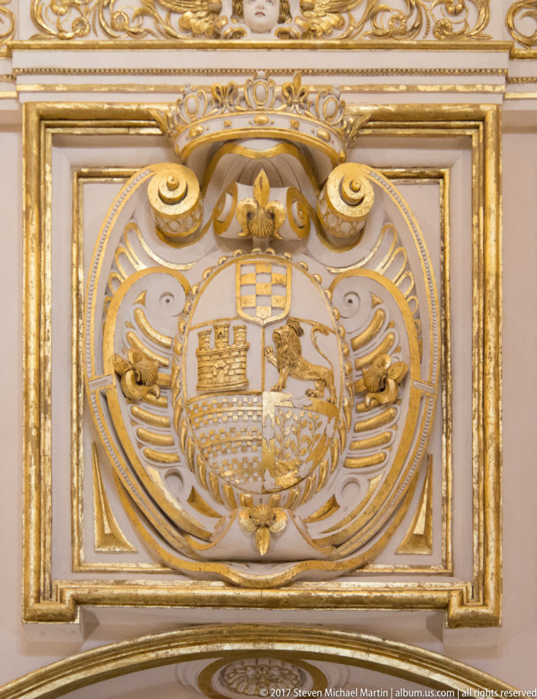 Gilded quartered coat of arms with castle and lion motifs framed by symmetrical scrollwork.