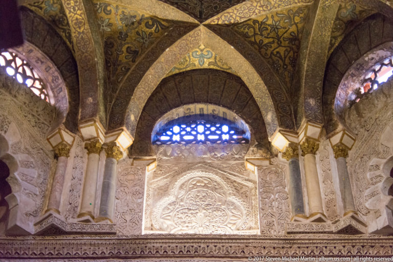 Pointed decorated vault with gold vegetal patterns above carved stonework and a blue-lit traceried opening.